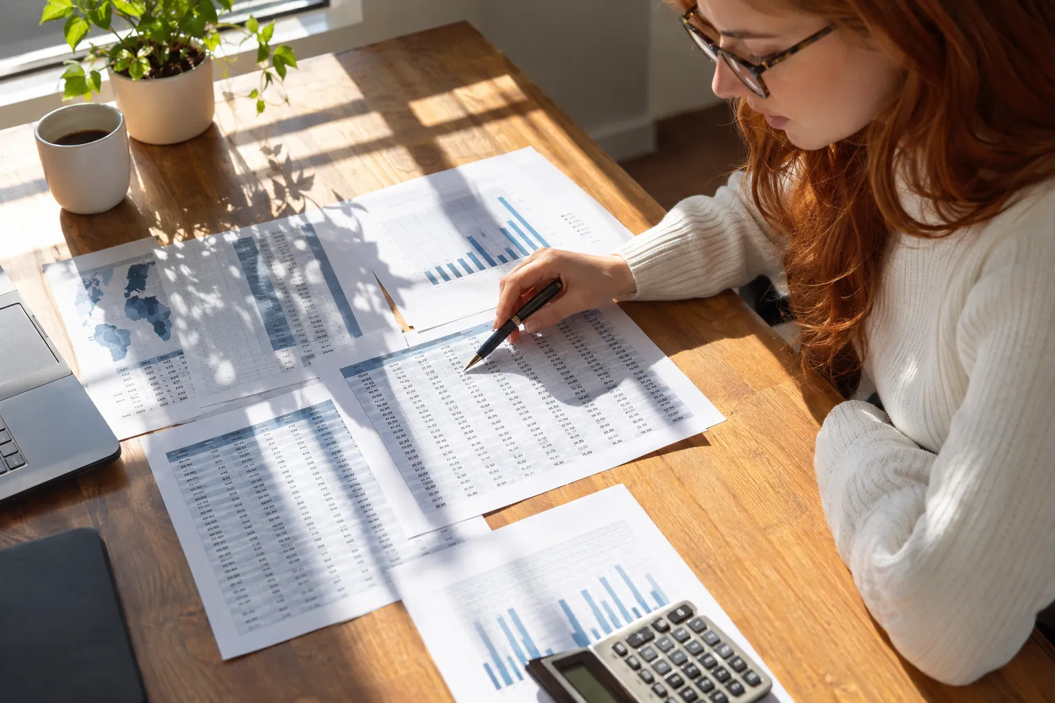 Femme travaillant sur des documents financiers et graphiques sur un bureau en bois, avec calculatrice et plantes en arrière-plan. travailler dans un hôpital français à l'étranger prête à être exploré (non visible mais lié).